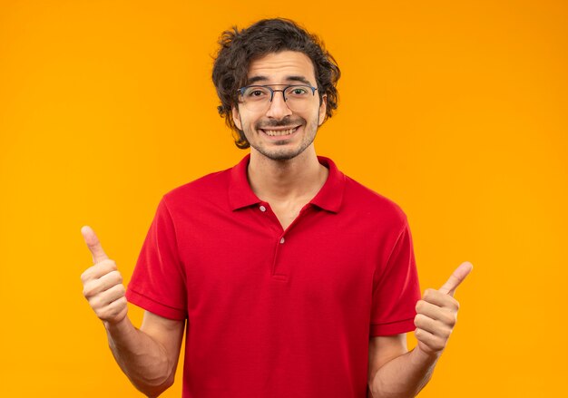 Young smiling man in red shirt with optical glasses thumbs up isolated on orange wall