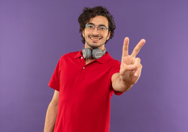 Young smiling man in red shirt with optical glasses and with headphones gestures victory hand sign isolated on violet wall