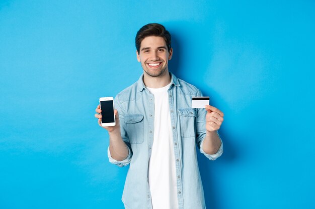 Young smiling man showing smartphone screen and credit card, concept of online shopping or banking.