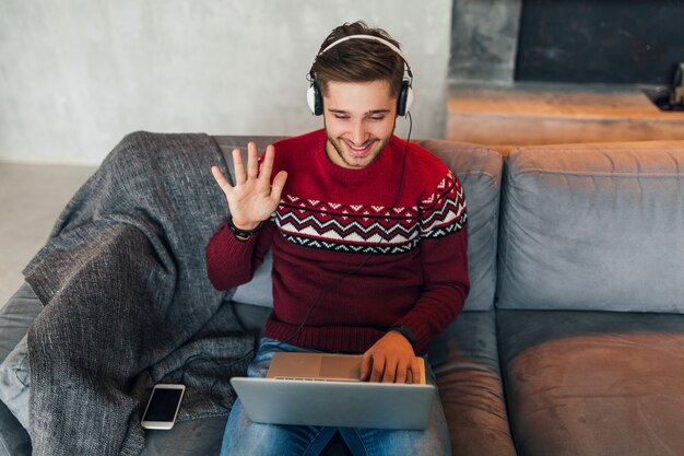Молодой улыбающийся человек сидит дома зимой, разговаривает в Интернете, машет рукой, здоровается, одет в красный свитер, работает на ноутбуке, фрилансер, слушает наушники, учится онлайн