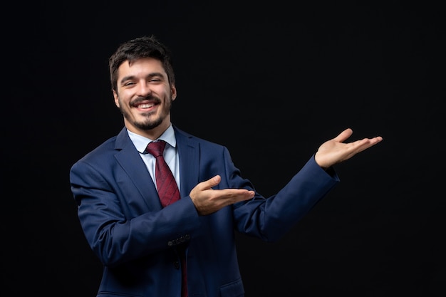 Young smiling man in suit pointing something on the left side on isolated dark wall