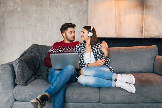 Young smiling man and woman sitting at home in winter, working on laptop, holding smartphone, listening to headphones, couple on leisure spending time together, freelancer, happy, dating