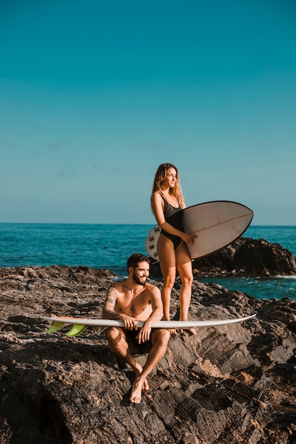 바다 근처 바위에 서핑 보드와 함께 젊은 웃는 남자와 여자