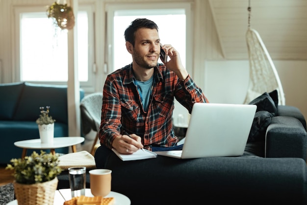 Young smiling man writing notes while making a phone call and using laptop at home