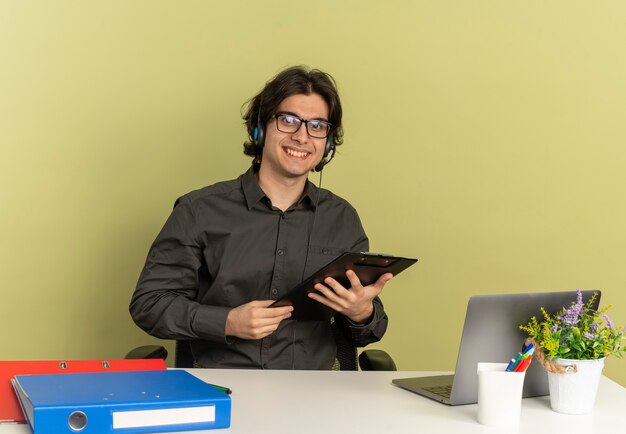 Giovane sorridente lavoratore di ufficio uomo sulle cuffie in vetri ottici si siede alla scrivania con strumenti per ufficio utilizzando laptop tenendo appunti isolato su sfondo verde con spazio di copia