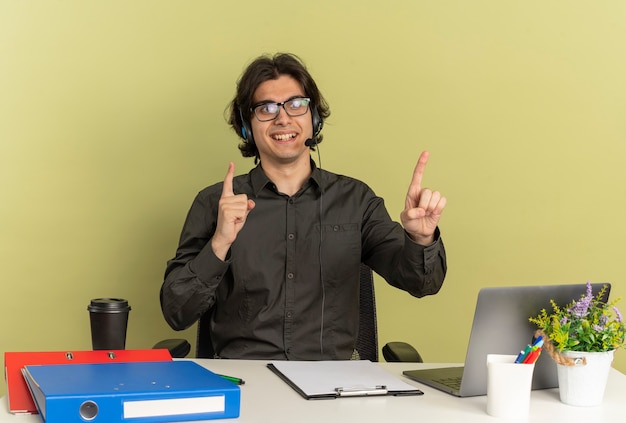 Young smiling office worker man on headphones in optical glasses sits at desk with office tools using laptop points up isolated on green background with copy space