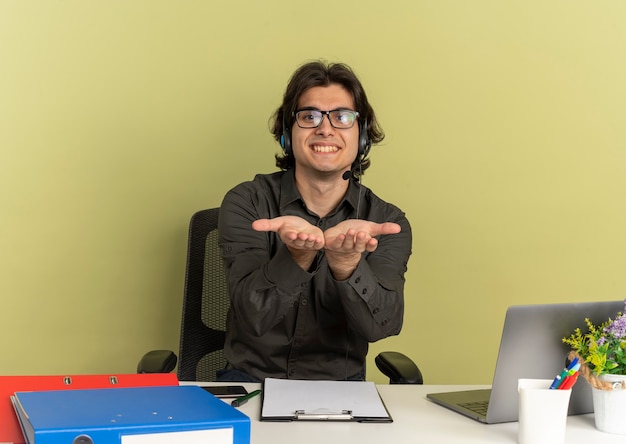 Free photo young smiling office worker man  in optical glasses sits at desk with office tools using laptop and holds hands open looking at camera isolated on green background with copy space