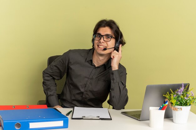 Young smiling office worker man in optical glasses sits at desk with office tools using laptop puts hand on headphones isolated on green background with copy space