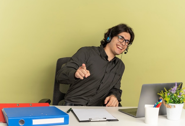 Free photo young smiling office worker manon headphones in optical glasses sits at desk with office tools using laptop pointing at camera isolated on green background with copy space