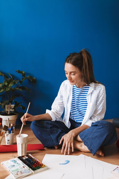 Young smiling painter in white shirt and striped T-shirt sitting on floor dreamily drawing while spending time at cozy home