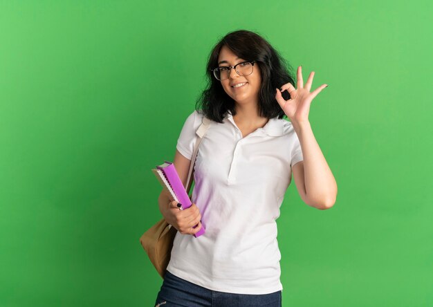 Young smiling pretty caucasian schoolgirl wearing glasses and back bag gestures ok hand sign holding books on green  with copy space