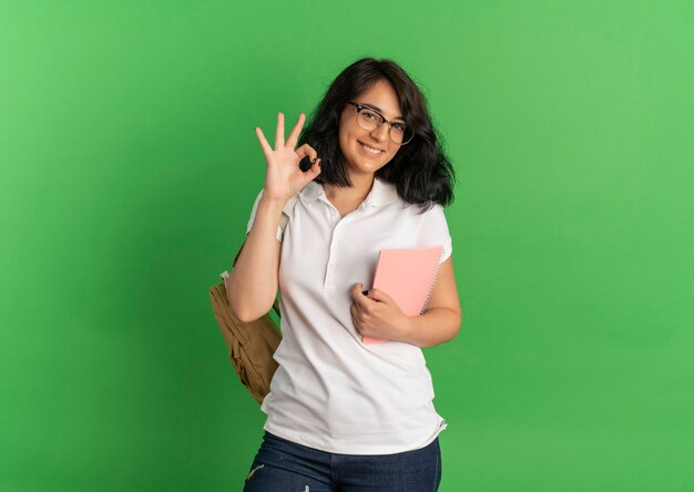 Молодая улыбающаяся симпатичная кавказская школьница в очках и жестах на спине, рука знак, держащая ноутбук на зеленом с копией пространства