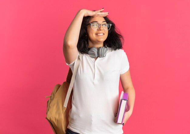 Young smiling pretty caucasian schoolgirl wearing glasses back bag and headphones on neck keeping palm at her forehead trying to see something holding book and notebook
