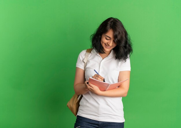 Young smiling pretty caucasian schoolgirl wearing glasses and back bag holds pen writes in notebook on green  with copy space