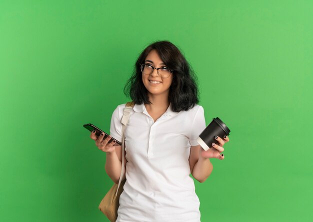 Young smiling pretty caucasian schoolgirl wearing glasses and back bag holds phone and coffee cup looking at side on green  with copy space