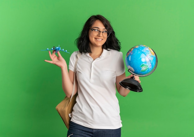 Young smiling pretty caucasian schoolgirl wearing glasses and back bag holds toy plane and globe on green  with copy space