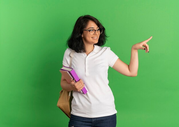 Young smiling pretty caucasian schoolgirl wearing glasses and back bag looks and points at side holding books on green  with copy space