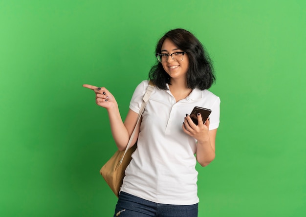 Young smiling pretty caucasian schoolgirl wearing glasses and back bag points at side holding phone on green  with copy space