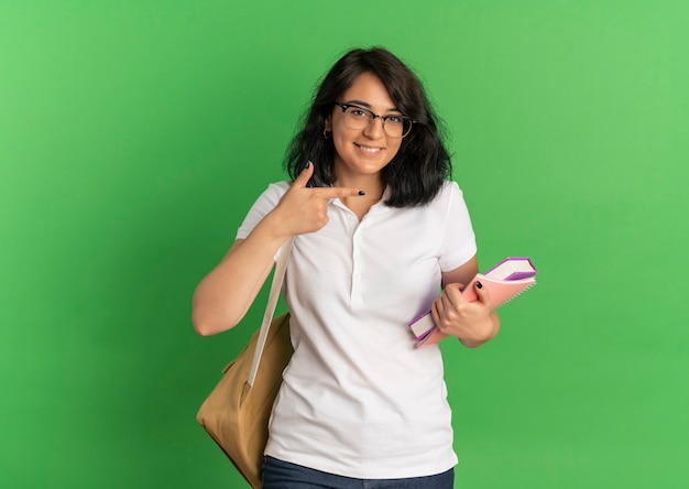 Young smiling pretty caucasian schoolgirl wearing glasses and back bag points at side and holds books on green  with copy space