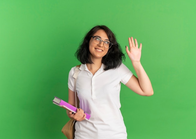 Young smiling pretty caucasian schoolgirl wearing glasses and back bag raises hand up looking at side holding books on green  with copy space