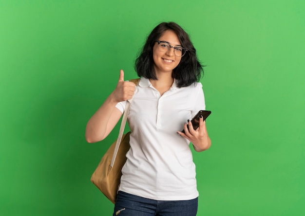 Young smiling pretty caucasian schoolgirl wearing glasses and back bag thumbs up holding phone on green  with copy space