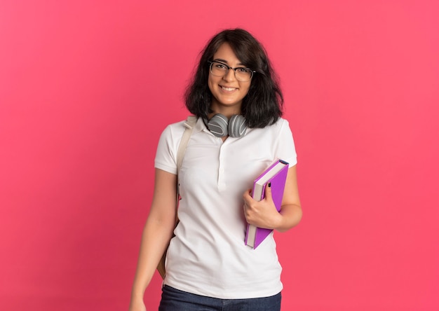 Молодая улыбающаяся симпатичная кавказская школьница в очках и задней сумке с наушниками на шее стоит, держа книги на розовом с копией пространства