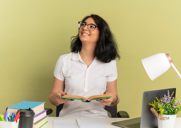 眼鏡をかけている若い笑顔のかなり白人の女子高生は、学校のツールで机に座ってコピースペースで緑の本を持って見上げる