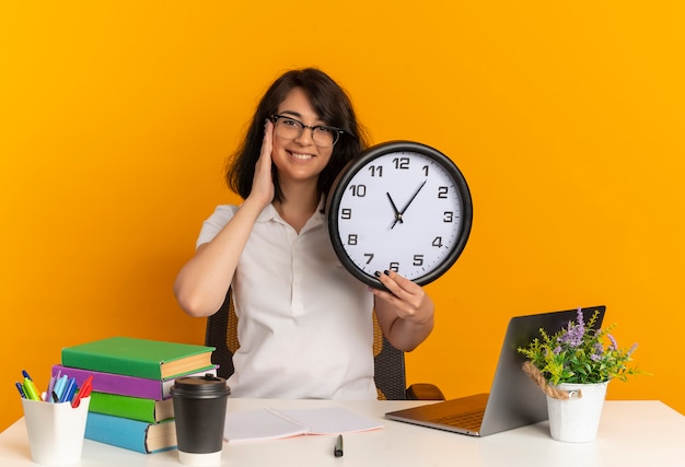 Young smiling pretty caucasian schoolgirl wearing glasses sits at desk with school tools puts hand on face holds clock isolated on orange space with copy space