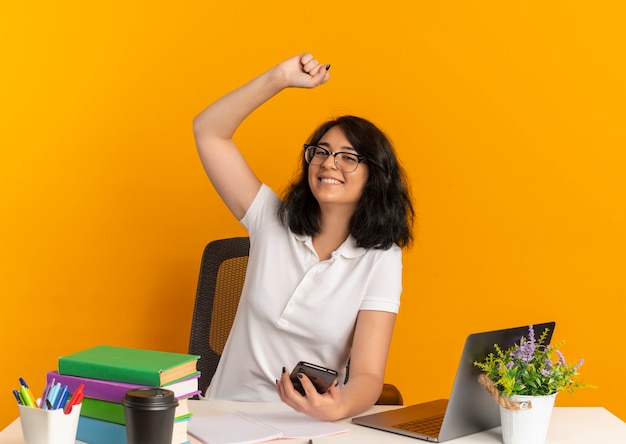 Young smiling pretty caucasian schoolgirl wearing glasses sits at desk with school tools raises fist up holds phone on orange  with copy space