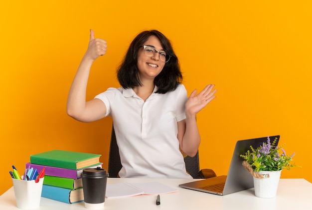Young smiling pretty caucasian schoolgirl wearing glasses sits at desk with school tools thumbs up and points with hand at side isolated on orange space with copy space