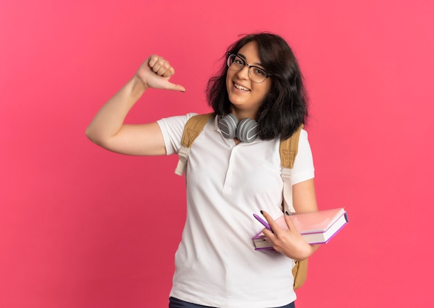 Young smiling pretty caucasian schoolgirl with headphones on neck wearing glasses and back bag points at herself holding books on pink  with copy space