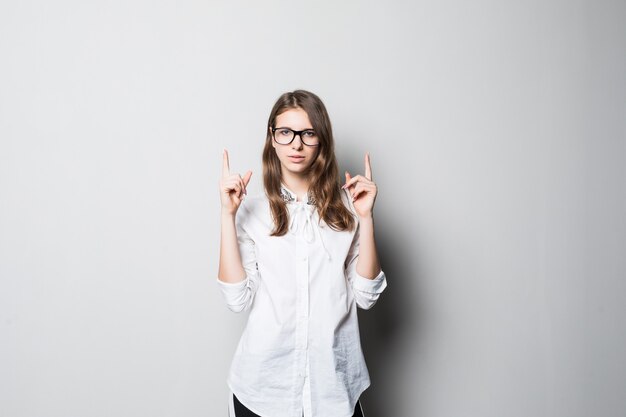 Young smiling pretty girl in glasses dressed up in strict office white t-shirt stands in front of white wall