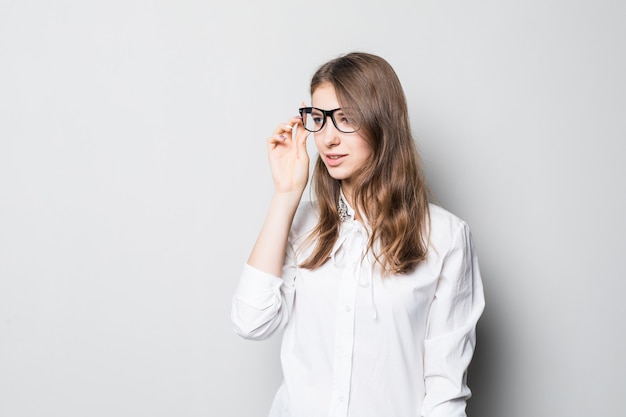 Young smiling pretty girl in glasses dressed up in strict office white t-shirt stands in front of white wall