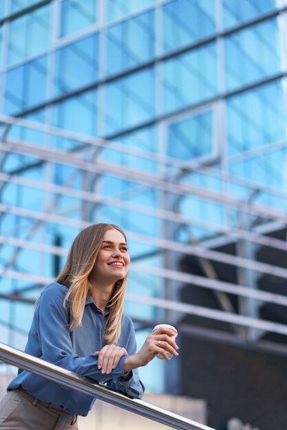 彼女の完全な稼働日の間にコーヒーブレークを持つ若い笑顔専門職の女性。彼女はリラックスして彼女の飲み物を楽しんでいる間、ビジネスビルの近くの屋外に紙コップを持っています。