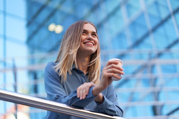 彼女の完全な稼働日の間にコーヒーブレークを持つ若い笑顔専門職の女性。彼女はリラックスして彼女の飲み物を楽しんでいる間、ビジネスビルの近くの屋外に紙コップを持っています。