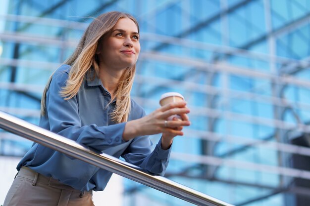 Young smiling professional woman having a coffee break during her full working day. She holds a paper cup outdoors near the business building while relaxing and enjoying her beverage.