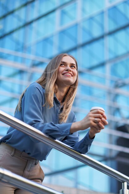 Young smiling professional woman having a coffee break during her full working day. She holds a paper cup outdoors near the business building while relaxing and enjoying her beverage.