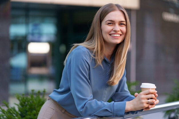 彼女の完全な稼働日の間にコーヒーブレークを持つ若い笑顔専門職の女性。彼女はリラックスして彼女の飲み物を楽しんでいる間、ビジネスビルの近くの屋外に紙コップを持っています。