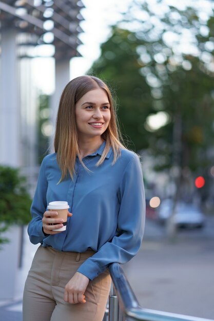 彼女の完全な稼働日の間にコーヒーブレークを持つ若い笑顔専門職の女性。彼女はリラックスして彼女の飲み物を楽しんでいる間、ビジネスビルの近くの屋外に紙コップを持っています。