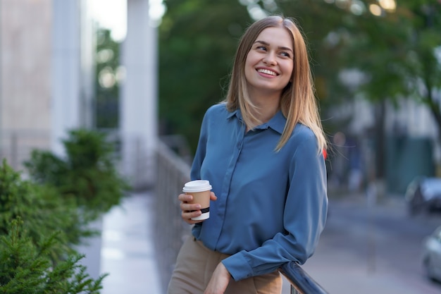 彼女の完全な稼働日の間にコーヒーブレークを持つ若い笑顔専門職の女性。彼女はリラックスして彼女の飲み物を楽しんでいる間、ビジネスビルの近くの屋外に紙コップを持っています。