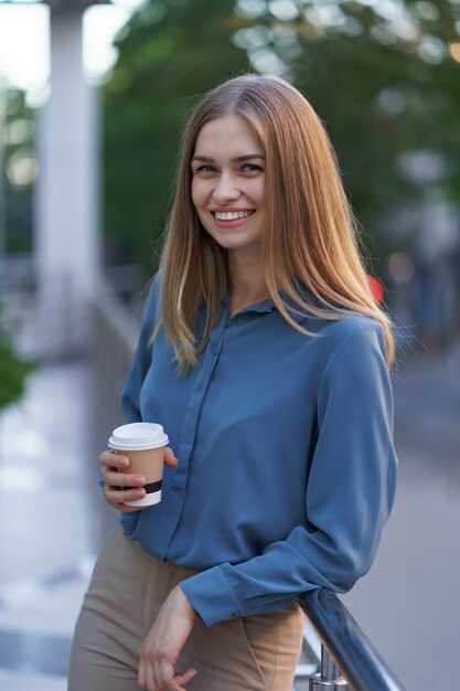 彼女の完全な稼働日の間にコーヒーブレークを持つ若い笑顔専門職の女性。彼女はリラックスして彼女の飲み物を楽しんでいる間、ビジネスビルの近くの屋外に紙コップを持っています。