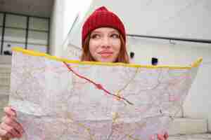 Foto gratuita il giovane turista sorridente della ragazza della testarossa si siede sulle scale all'aperto con la mappa di carta della città che cerca la direzione