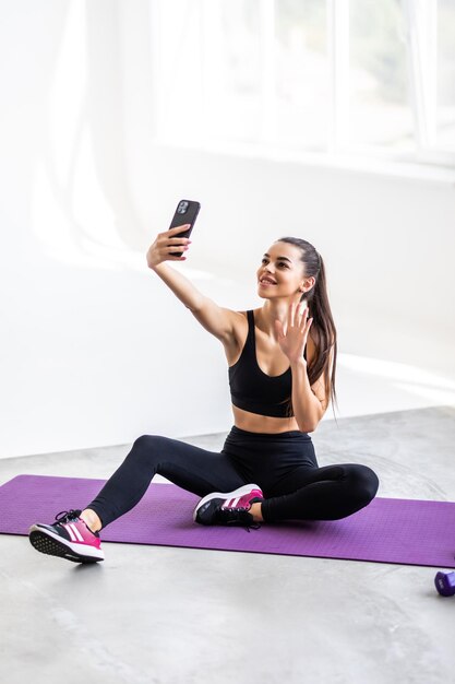 Young smiling strong sporty athletic fitness trainer instructor woman doing exercises do selfie shot on mobile cell phone sit on mat floor at home gym