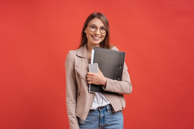 Молодой усмехаясь студент или интерн в eyeglasses стоя с папкой на красной стене.
