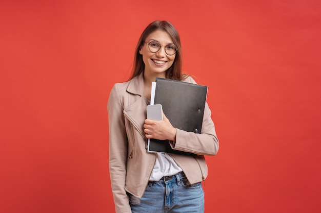 Young smiling student or intern in eyeglasses standing with a folder on red wall.