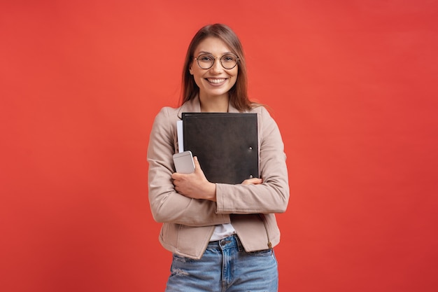 Free photo young smiling student or intern in eyeglasses standing with a folder on red wall.