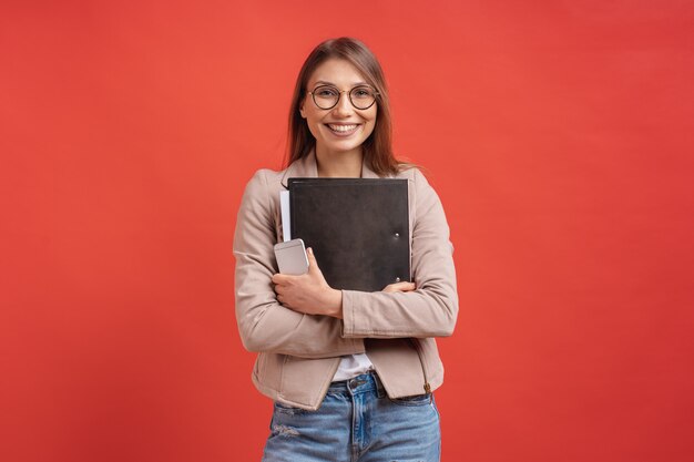 Young smiling student or intern in eyeglasses standing with a folder on red wall.