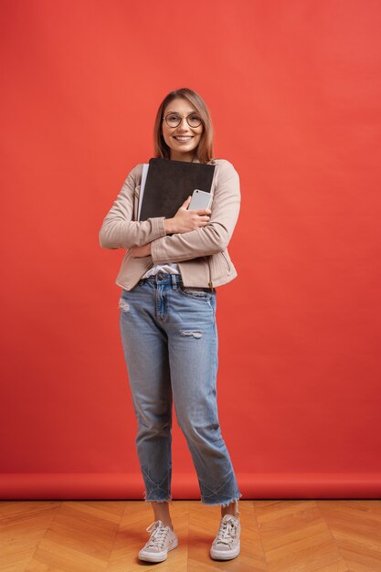 Young smiling student or intern in eyeglasses standing with a folder on red wall.