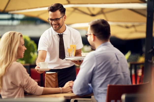 屋外カフェでカップルにコーヒーをブライニング若い笑顔のウェイター