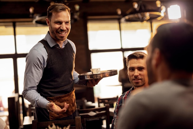 Free photo young smiling waiter serving male guests and talking to them in coffee shop